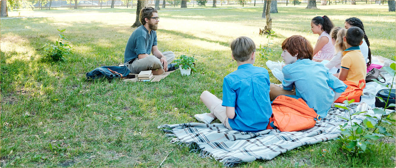 Young bearded botanist in eyeglasses sitting with potted plant on ground and teaching pupils in park